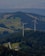 Two large wind turbines are situated in a lush, forested area with hills and scattered clearings. A small road winds through the greenery, leading to a building nestled among the trees. The landscape features expansive views of rolling hills, fields, and forested areas.