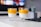 Two glasses of chia pudding topped with a bright orange mango puree are placed on a white table. The background includes a blurred television screen and a small potted plant.