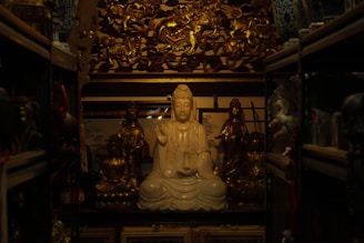 A serene, white statue of a meditative figure seated in the center, surrounded by ornate golden statues and intricate carvings. The setting is dimly lit, creating a warm, contemplative atmosphere with shelves containing various artifacts on either side.