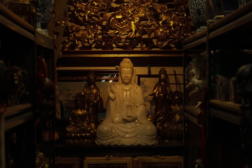 A serene, white statue of a meditative figure seated in the center, surrounded by ornate golden statues and intricate carvings. The setting is dimly lit, creating a warm, contemplative atmosphere with shelves containing various artifacts on either side.