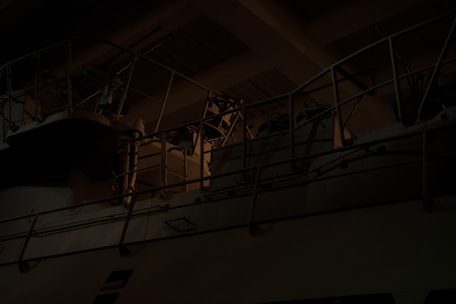 A dark, industrial setting featuring metal railings and possibly machinery with dim lighting creating shadows and highlights.