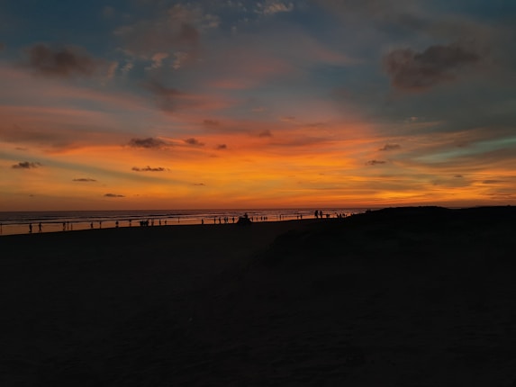 A breathtaking sunset over a beach with vibrant orange, yellow, and blue hues in the sky. Silhouettes of people are visible on the horizon, suggesting a serene evening scene.