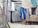 Clothing is hanging on a line in front of an old, rustic wooden door with weathered stone walls. A mix of garments, including a pair of jeans and shirts, sway slightly in the breeze. The setting has a vintage, rural feel with hints of neglect.