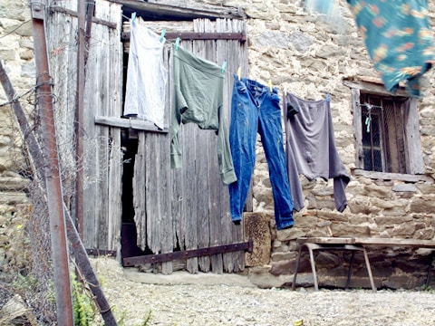 Clothing is hanging on a line in front of an old, rustic wooden door with weathered stone walls. A mix of garments, including a pair of jeans and shirts, sway slightly in the breeze. The setting has a vintage, rural feel with hints of neglect.