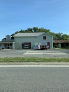 A small, light-colored building with signs for 'Benelli', 'Franchi', and 'Marlboro' on the front. There is one car parked in front, along with an ice vending machine. The building is surrounded by trees and there is a paved road in the foreground.