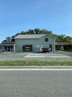 A small, light-colored building with signs for 'Benelli', 'Franchi', and 'Marlboro' on the front. There is one car parked in front, along with an ice vending machine. The building is surrounded by trees and there is a paved road in the foreground.