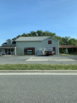 A small, light-colored building with signs for 'Benelli', 'Franchi', and 'Marlboro' on the front. There is one car parked in front, along with an ice vending machine. The building is surrounded by trees and there is a paved road in the foreground.