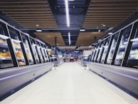 A spacious, brightly lit supermarket aisle with two rows of refrigerated display cases on either side. The floor is glossy and tiled, reflecting the overhead lighting. The ceiling has a modern design with wooden panels and exposed black ductwork. Various products can be seen inside the glass-fronted fridges along both sides. In the distance, a few people are visible, partially obscured by the displays.