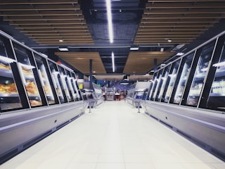 A spacious, brightly lit supermarket aisle with two rows of refrigerated display cases on either side. The floor is glossy and tiled, reflecting the overhead lighting. The ceiling has a modern design with wooden panels and exposed black ductwork. Various products can be seen inside the glass-fronted fridges along both sides. In the distance, a few people are visible, partially obscured by the displays.