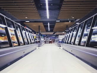 A spacious, brightly lit supermarket aisle with two rows of refrigerated display cases on either side. The floor is glossy and tiled, reflecting the overhead lighting. The ceiling has a modern design with wooden panels and exposed black ductwork. Various products can be seen inside the glass-fronted fridges along both sides. In the distance, a few people are visible, partially obscured by the displays.