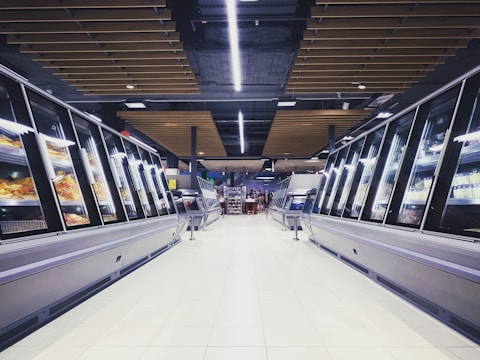 A spacious, brightly lit supermarket aisle with two rows of refrigerated display cases on either side. The floor is glossy and tiled, reflecting the overhead lighting. The ceiling has a modern design with wooden panels and exposed black ductwork. Various products can be seen inside the glass-fronted fridges along both sides. In the distance, a few people are visible, partially obscured by the displays.