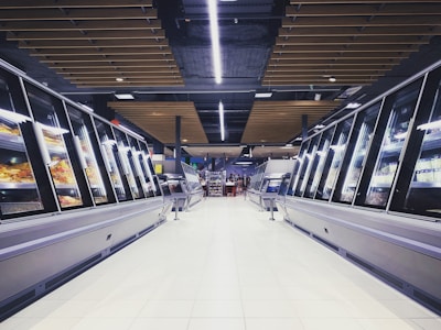 A spacious, brightly lit supermarket aisle with two rows of refrigerated display cases on either side. The floor is glossy and tiled, reflecting the overhead lighting. The ceiling has a modern design with wooden panels and exposed black ductwork. Various products can be seen inside the glass-fronted fridges along both sides. In the distance, a few people are visible, partially obscured by the displays.