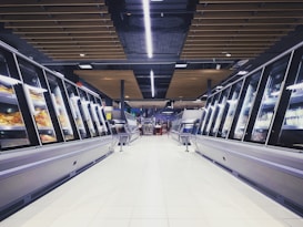 A spacious, brightly lit supermarket aisle with two rows of refrigerated display cases on either side. The floor is glossy and tiled, reflecting the overhead lighting. The ceiling has a modern design with wooden panels and exposed black ductwork. Various products can be seen inside the glass-fronted fridges along both sides. In the distance, a few people are visible, partially obscured by the displays.