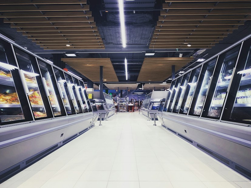 A spacious, brightly lit supermarket aisle with two rows of refrigerated display cases on either side. The floor is glossy and tiled, reflecting the overhead lighting. The ceiling has a modern design with wooden panels and exposed black ductwork. Various products can be seen inside the glass-fronted fridges along both sides. In the distance, a few people are visible, partially obscured by the displays.