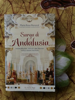 A book titled 'Surga di Andalusia' by Maria Rosa Menocal, discussing the harmony among Muslims, Jews, and Christians in Andalusia. The cover features intricate Islamic architectural designs and an illustration of historical figures engaging in dialogue. The background consists of decorative patterns.