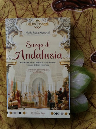 A book titled 'Surga di Andalusia' by Maria Rosa Menocal, discussing the harmony among Muslims, Jews, and Christians in Andalusia. The cover features intricate Islamic architectural designs and an illustration of historical figures engaging in dialogue. The background consists of decorative patterns.