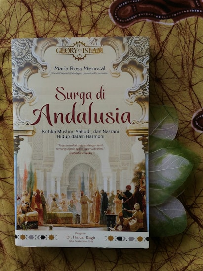 A book titled 'Surga di Andalusia' by Maria Rosa Menocal, discussing the harmony among Muslims, Jews, and Christians in Andalusia. The cover features intricate Islamic architectural designs and an illustration of historical figures engaging in dialogue. The background consists of decorative patterns.