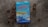 A book titled 'Misteri Soliter' by Jostein Gaarder is placed on a wooden surface surrounded by dried brown leaves. The book cover is predominantly blue with red and yellow text. It features an illustration of architectural structures and a sky with balloons.