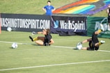 Two football players are performing a training drill on a grassy field. One is leaning back while controlling a soccer ball with her hands, and the other is lying on her back with her legs and arms up, holding another soccer ball. Both are wearing black attire with yellow socks. In the background, banners display social media handles and web addresses, and a person in a blue shirt stands nearby, watching the scene.