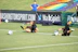 Two football players are performing a training drill on a grassy field. One is leaning back while controlling a soccer ball with her hands, and the other is lying on her back with her legs and arms up, holding another soccer ball. Both are wearing black attire with yellow socks. In the background, banners display social media handles and web addresses, and a person in a blue shirt stands nearby, watching the scene.