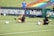 Two football players are performing a training drill on a grassy field. One is leaning back while controlling a soccer ball with her hands, and the other is lying on her back with her legs and arms up, holding another soccer ball. Both are wearing black attire with yellow socks. In the background, banners display social media handles and web addresses, and a person in a blue shirt stands nearby, watching the scene.