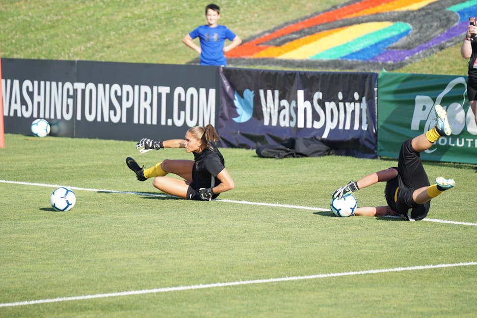 Two football players are performing a training drill on a grassy field. One is leaning back while controlling a soccer ball with her hands, and the other is lying on her back with her legs and arms up, holding another soccer ball. Both are wearing black attire with yellow socks. In the background, banners display social media handles and web addresses, and a person in a blue shirt stands nearby, watching the scene.
