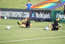 Two football players are performing a training drill on a grassy field. One is leaning back while controlling a soccer ball with her hands, and the other is lying on her back with her legs and arms up, holding another soccer ball. Both are wearing black attire with yellow socks. In the background, banners display social media handles and web addresses, and a person in a blue shirt stands nearby, watching the scene.
