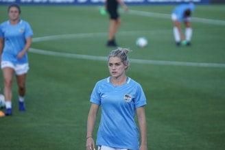 A woman in a light blue soccer jersey with a sports logo stands on a green soccer field. In the background, other players in similar uniforms are visible, with one stretching and another in motion. A soccer ball is also seen in the distance.