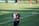 A person wearing black sports attire and a towel draped over their shoulder stands on a grassy sports field. In the background, there are other athletes and a banner with partially visible text.