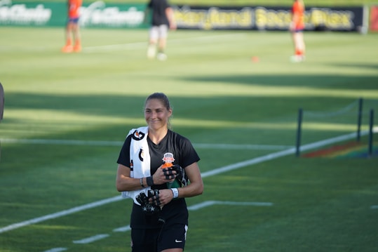 A person wearing black sports attire and a towel draped over their shoulder stands on a grassy sports field. In the background, there are other athletes and a banner with partially visible text.