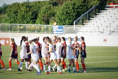 Players from two soccer teams are shaking hands on the field. They are wearing sports uniforms, one team in white and the other in dark uniforms with red accents. The setting appears to be a soccer field with green grass, and there are bleachers visible in the background.