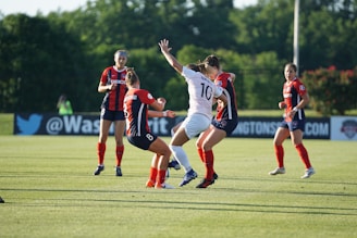 A group of women soccer players actively engaged in the game on a green soccer field. Three players in red and black uniforms are interacting with a player in a white uniform. The background features trees and some banners along the side of the field, indicating a professional soccer match environment.
