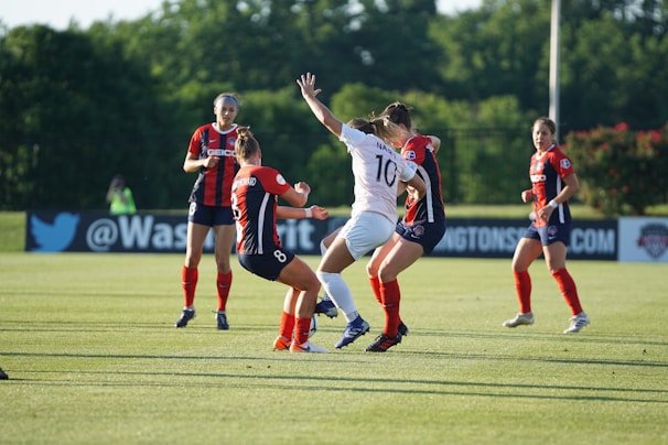 A group of women soccer players actively engaged in the game on a green soccer field. Three players in red and black uniforms are interacting with a player in a white uniform. The background features trees and some banners along the side of the field, indicating a professional soccer match environment.