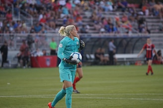 A soccer player in a turquoise uniform is holding a soccer ball on a grassy field. The player appears to be focused on the game, with stands full of spectators blurred in the background. Another player in a red and blue uniform can be seen in the background.