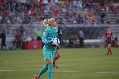 A soccer player in a turquoise uniform is holding a soccer ball on a grassy field. The player appears to be focused on the game, with stands full of spectators blurred in the background. Another player in a red and blue uniform can be seen in the background.