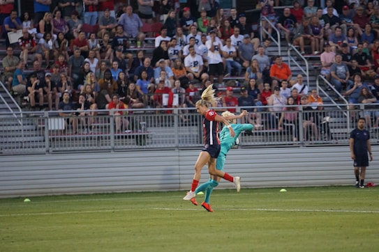 Two soccer players, one in a dark uniform with red accents and the other in a teal uniform, are engaged in a dynamic action on the field. The players are caught mid-action with an audience in the background seated in stadium bleachers.