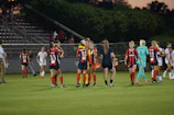 A group of female soccer players are on the field, some wearing red and navy jerseys, while others wear white jerseys. They are interacting casually, with some players holding drinks, and a coach carrying orange Gatorade containers.