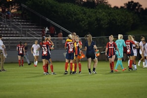 A group of female soccer players are on the field, some wearing red and navy jerseys, while others wear white jerseys. They are interacting casually, with some players holding drinks, and a coach carrying orange Gatorade containers.