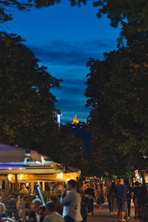 A bustling street scene at dusk features a lively outdoor dining area with people socializing and enjoying their evening. The sky is a deep blue, and trees frame the view, leading the eye towards a brightly lit structure on a distant hilltop. The warm glow from the dining area contrasts with the cool tones of the evening sky.