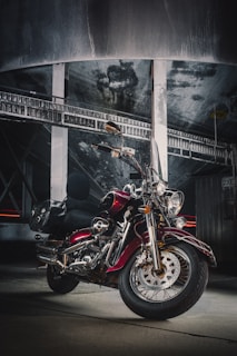 A classic motorcycle with a shiny red finish and chrome detailing is parked indoors. The setting has an industrial vibe with a concrete floor and a large metallic structure in the background. The lighting is dramatic, highlighting the contours and features of the motorcycle.
