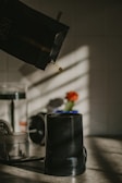 A coffee bean is being poured from a dark-colored bag into a black coffee grinder. The scene is set on a countertop with soft natural light casting shadows. In the background, there's a blurry orange flower in a vase and other kitchen objects.