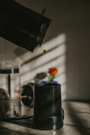 A coffee bean is being poured from a dark-colored bag into a black coffee grinder. The scene is set on a countertop with soft natural light casting shadows. In the background, there's a blurry orange flower in a vase and other kitchen objects.