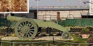 A large, historical-looking cannon displayed in an outdoor area, surrounded by manicured grass and small bushes. The cannon is painted green with detailed wheels and sits on a bed of rocks. Metal railings and decorative elements are visible in the background, as well as a section of a building with green fencing and billboards.