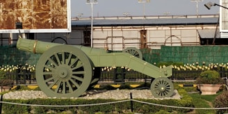 A large, historical-looking cannon displayed in an outdoor area, surrounded by manicured grass and small bushes. The cannon is painted green with detailed wheels and sits on a bed of rocks. Metal railings and decorative elements are visible in the background, as well as a section of a building with green fencing and billboards.