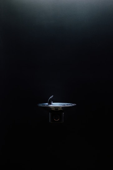 A drinking fountain is centered in an otherwise dimly lit environment, with light highlighting its metallic surface against the dark background.