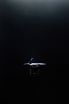 A drinking fountain is centered in an otherwise dimly lit environment, with light highlighting its metallic surface against the dark background.