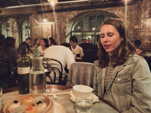 A woman in a denim jacket with long hair sits at a restaurant table with dim lighting. In front of her are a bowl, a glass, and a plate of Asian dumplings. The setting includes a rustic brick interior with several other diners in the background, creating a cozy and bustling atmosphere.