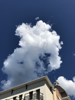 A white cloud with a fluffy texture floats against a deep blue sky. The corner of a building with a white, light brown, and gray facade is visible at the bottom, featuring black metal railings and a small, cylindrical turret.