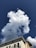 A white cloud with a fluffy texture floats against a deep blue sky. The corner of a building with a white, light brown, and gray facade is visible at the bottom, featuring black metal railings and a small, cylindrical turret.