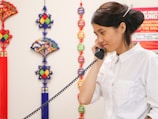 A person wearing a white shirt is speaking on a corded phone with a relaxed expression. The background features colorful decorative ornaments including tassels and fans, with a red and blue theme.